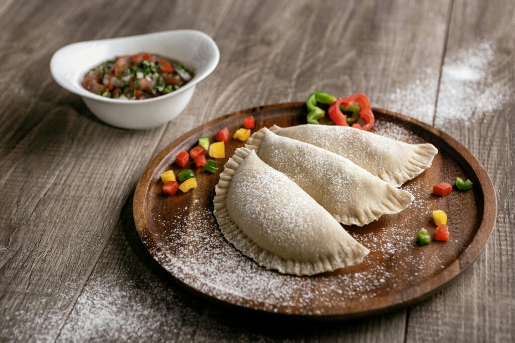 Three empanadas on a wooden plate with powdered sugar, served with dipping sauce on a wooden table.